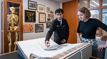 A library worker and a patron interact with a large book open to a skull diagram. A skeleton model and photographs are hung up on the wall.