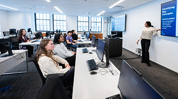 An instructor, pointing to a large screen next to her, teaches a class of students who sit at long tables with computers. 