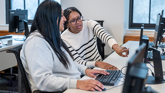 Seated at a long table filled with desktop computers, a librarian points at a patron's screen. The patron has her hands on the keyboard and looks engaged.