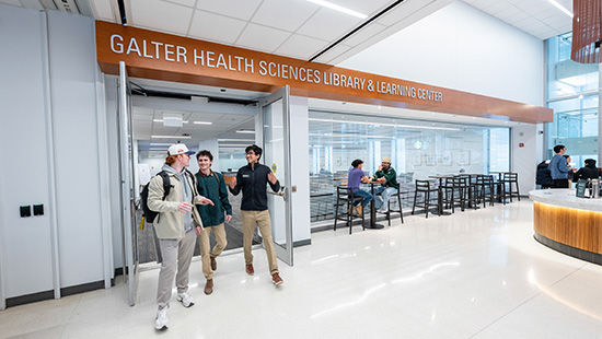 A group of three students exit the Galter Library. Other Feinberg community members are talking over coffee in the background.