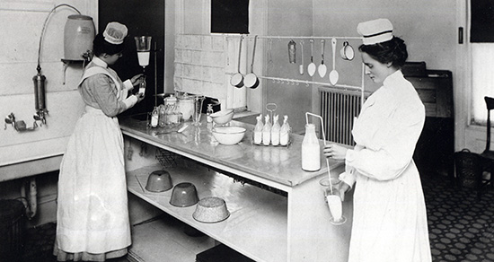 A black-and-white photo from circa 1906 showing two nurses in white uniforms and caps working in Northwestern’s diet kitchen. The nurse on the left stands near a sink filling small glass bottles, while the nurse on the right stands at a prep table handling a large milk bottle. The table is organized with racks of infant formula bottles, bowls, and hanging spoons