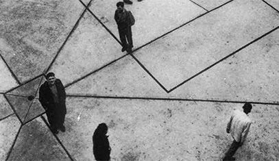 A black-and-white photograph of people standing apart on paved ground broken up by dark lines